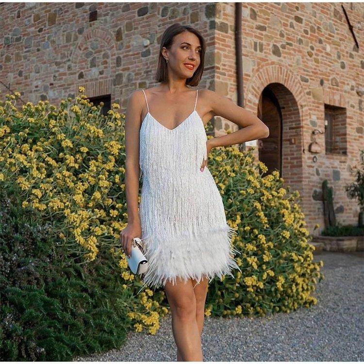 Woman in white fringe dress with spaghetti straps, holding clutch, standing by yellow flowers near brick building