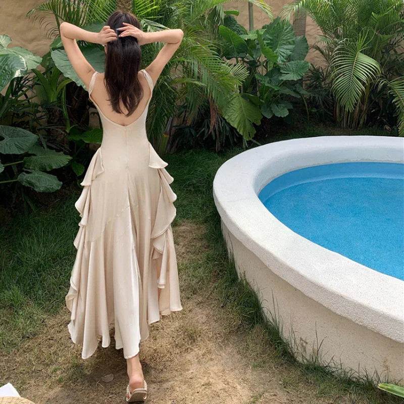 Woman in elegant beige maxi dress with ruffled layers, standing by poolside garden, showcasing backless design