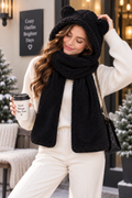 A woman wearing a cozy black sherpa hooded scarf with bear ears and a matching black fleece scarf, styled with a cream ribbed sweater and cream pants in an outdoor holiday setting.