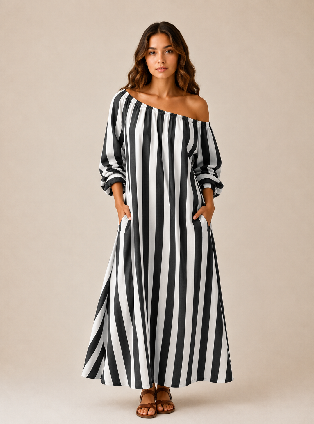A woman modeling a black and white vertically striped off-the-shoulder maxi dress with long sleeves and side pockets, standing against a solid beige studio background.