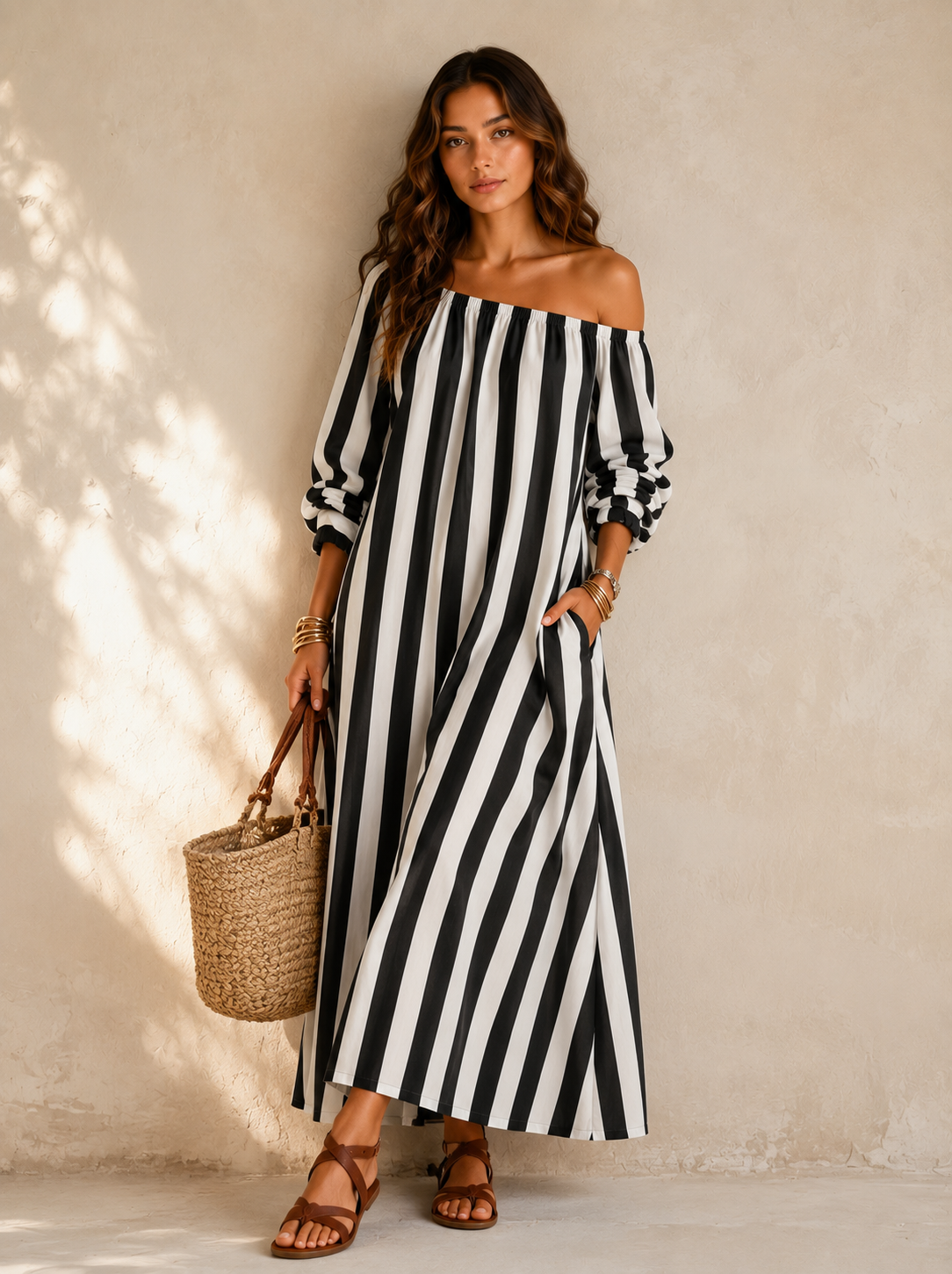 A woman models an off-the-shoulder maxi dress with bold black and white vertical stripes. She is holding a woven straw tote bag and wearing brown leather sandals, standing against a textured beige wall with soft leaf shadows.
