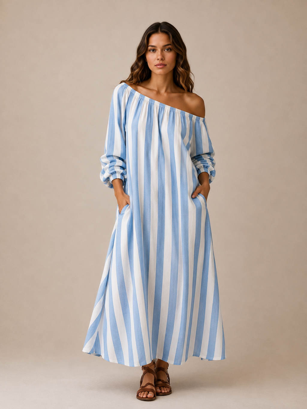 A woman modeling a light blue and white vertically striped off-the-shoulder maxi dress with long puff sleeves and side pockets, paired with brown strappy sandals, against a solid beige studio background.