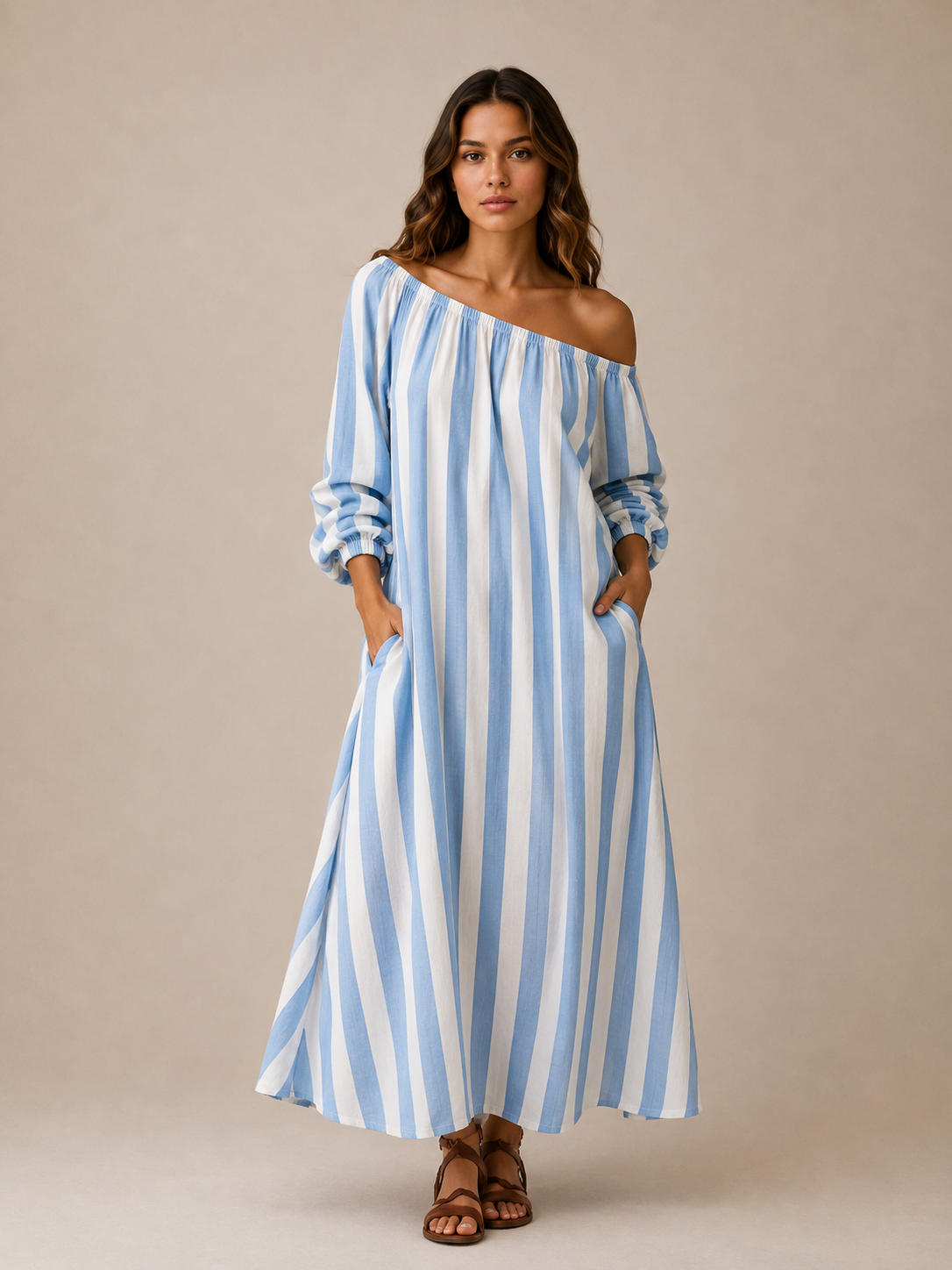 A woman modeling a light blue and white vertically striped off-the-shoulder maxi dress with long puff sleeves and side pockets, paired with brown strappy sandals, against a solid beige studio background.