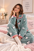 A woman sits on a bed wearing a cozy matching pajama set with a teal-green base, pink trim, and a white cartoon penguin print. The room is warmly lit with a lamp, framed wall art, and a pink pillow that reads 'Good Night'.