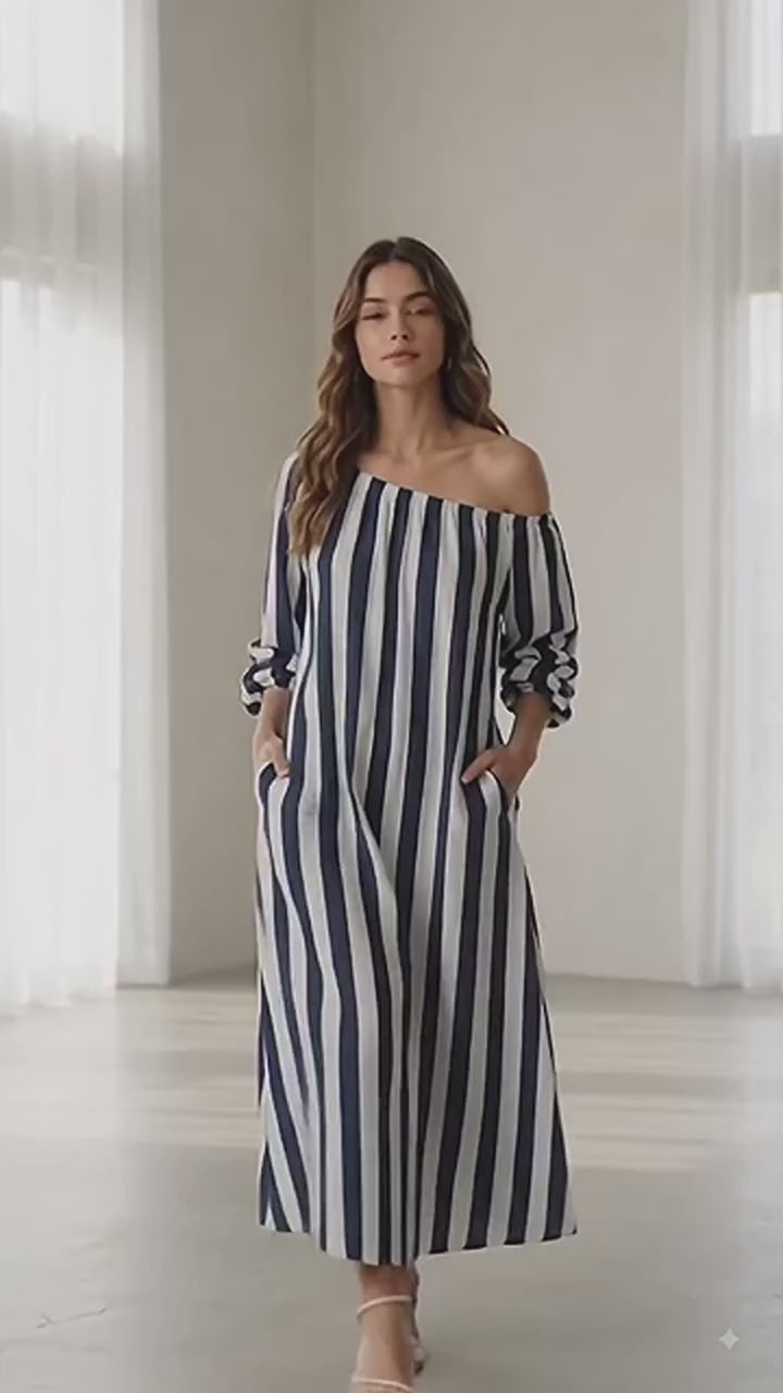 A woman modeling a long off-the-shoulder maxi dress with vertical burgundy and off-white stripes, walking against a plain beige studio background.